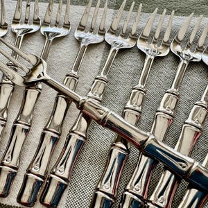 May include: A collection of silver-colored forks with bamboo-style handles. The forks are arranged on a light gray and white patterned surface, with the tines of the forks facing upwards.