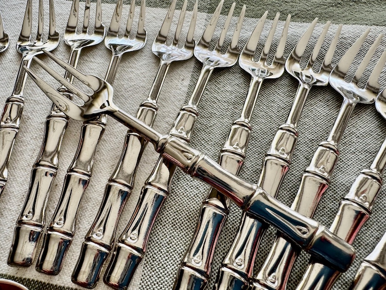 May include: A collection of silver-colored forks with bamboo-style handles. The forks are arranged on a light gray and white patterned surface, with the tines of the forks facing upwards.