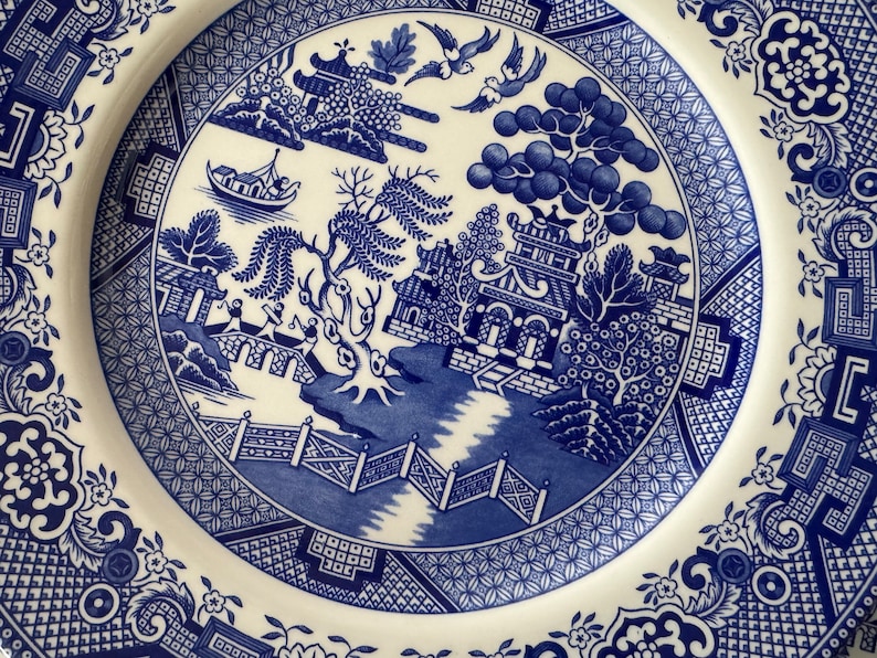 K&ouml;nnte beinhalten: Blau-wei&szlig;e Keramikplatte mit einem traditionellen chinesischen Weidenmuster. Das Design zeigt eine Pagode, eine Br&uuml;cke, einen Weidenbaum und einen Fluss.