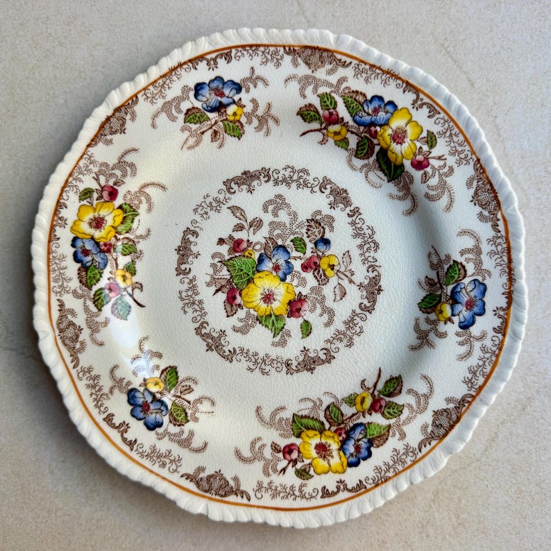May include: A round ceramic plate with a scalloped edge, featuring a cream-colored base decorated with brown scrollwork patterns and clusters of yellow and blue flowers with green leaves arranged in a central motif and around the rim.