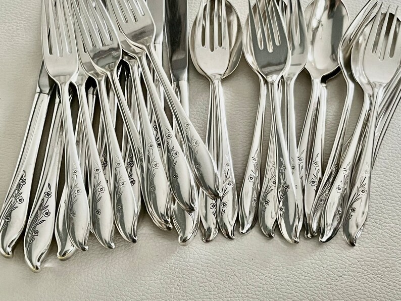May include: A collection of shiny silver flatware, including forks, spoons, and knives. Each piece is adorned with a delicate floral pattern along the handle. The silverware is arranged on a light-colored surface.