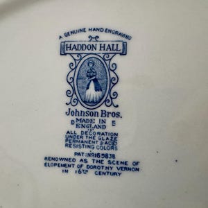 Op de afbeelding: Een wit keramisch oppervlak met een blauw gedrukt fabrieksmerk voor Haddon Hall door Johnson Bros. De tekst bevat A GENUINE HAND ENGRAVING, MADE IN ENGLAND, ALL DECORATION UNDER THE GLAZE en PAT NO 165838, met een centrale illustratie.
