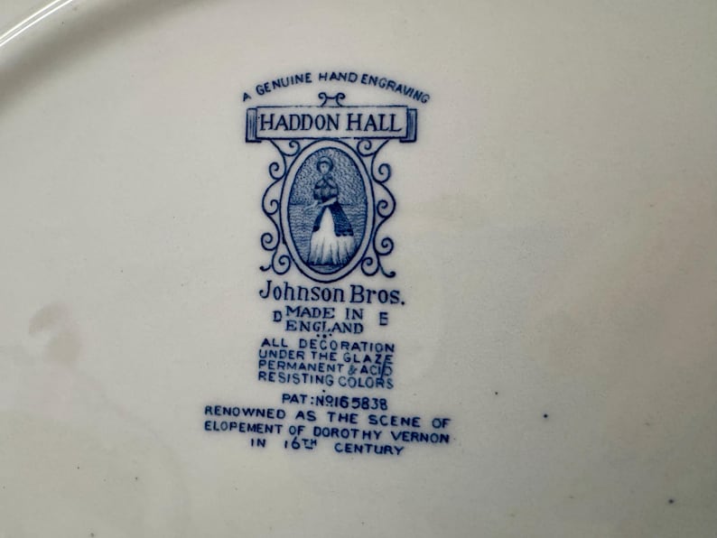Op de afbeelding: Een wit keramisch oppervlak met een blauw gedrukt fabrieksmerk voor Haddon Hall door Johnson Bros. De tekst bevat A GENUINE HAND ENGRAVING, MADE IN ENGLAND, ALL DECORATION UNDER THE GLAZE en PAT NO 165838, met een centrale illustratie.