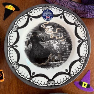 K&ouml;nnte beinhalten: Ein wei&szlig;er Keramik-Dinnerteller mit einer schwarz-wei&szlig;en Halloween-Szene, die eine Hexe zeigt, die auf einem Besen fliegt. Der Teller hat einen grauen Rand und einen schwarz-wei&szlig;en Rand mit Flederm&auml;usen und K&uuml;rbissen. Der Teller ist von Royal Stafford und tr&auml;gt die Aufschrift "Made in England".
