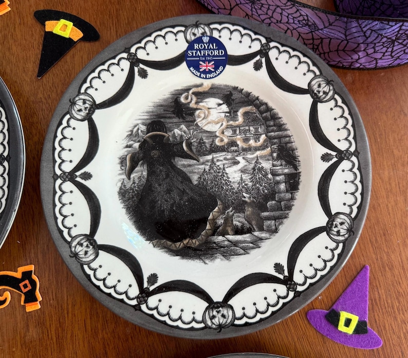 K&ouml;nnte beinhalten: Ein wei&szlig;er Keramik-Dinnerteller mit einer schwarz-wei&szlig;en Halloween-Szene, die eine Hexe zeigt, die auf einem Besen fliegt. Der Teller hat einen grauen Rand und einen schwarz-wei&szlig;en Rand mit Flederm&auml;usen und K&uuml;rbissen. Der Teller ist von Royal Stafford und tr&auml;gt die Aufschrift "Made in England".