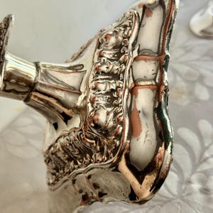 May include: A silver-plated candlestick base featuring an ornate, raised floral and scrollwork border. The metallic surface shows visible wear with copper-colored base metal showing through the silver finish along the edges and decorative relief.