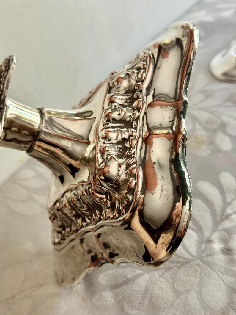 May include: A silver-plated candlestick base featuring an ornate, raised floral and scrollwork border. The metallic surface shows visible wear with copper-colored base metal showing through the silver finish along the edges and decorative relief.