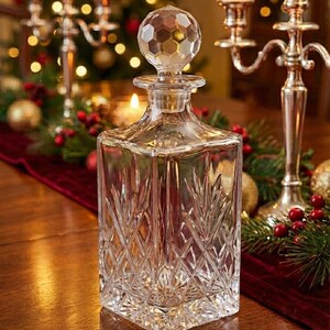 May include: A clear, square crystal decanter with a faceted stopper. The decanter features a diamond and leaf pattern. The background includes lit candles, a red runner, and Christmas decorations, suggesting a festive theme.