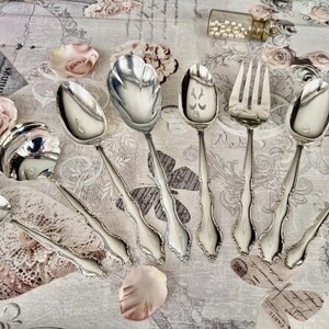 May include: A collection of ornate silver serving utensils, including spoons and a fork, arranged on a floral patterned surface. The utensils have detailed handles and decorative spoon bowls. The image also includes rose petals and a small glass vial.