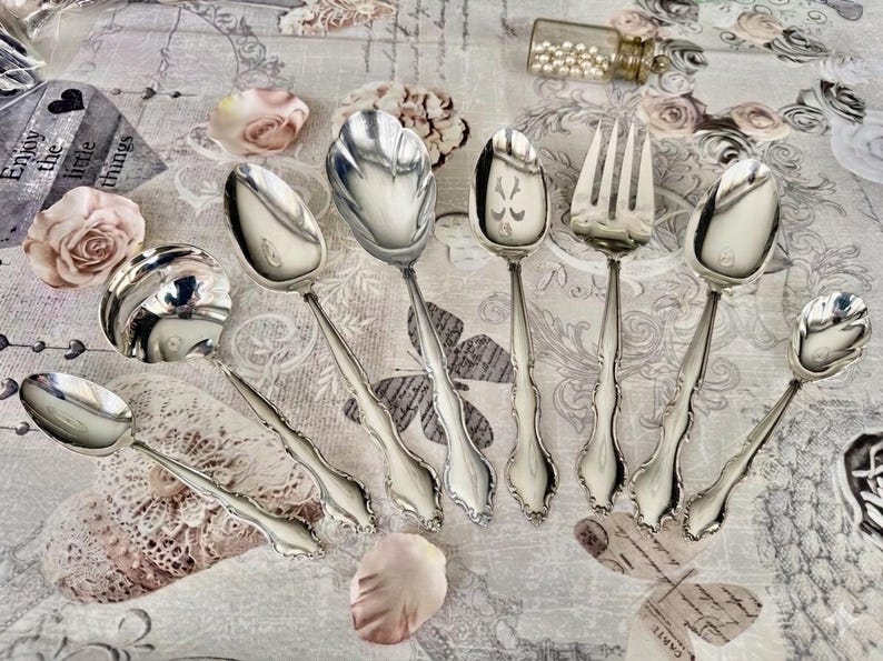May include: A collection of ornate silver serving utensils, including spoons and a fork, arranged on a floral patterned surface. The utensils have detailed handles and decorative spoon bowls. The image also includes rose petals and a small glass vial.
