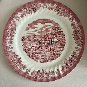 May include: A white ceramic dinner plate with a red and white floral design. The center of the plate features a cottage scene with a red roof and a watermill.