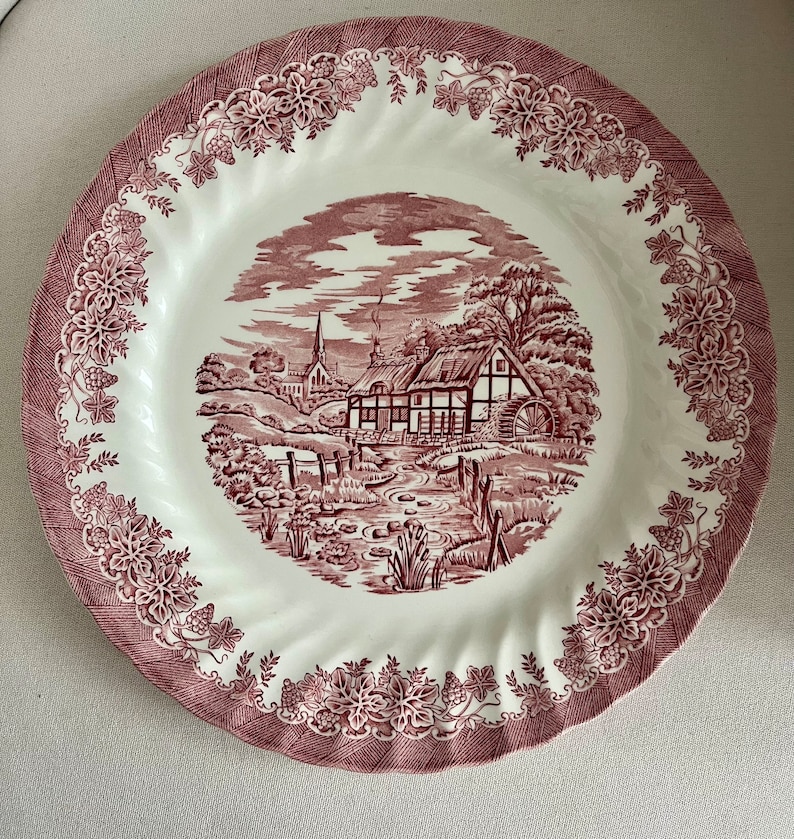 May include: A white ceramic dinner plate with a red and white floral design. The center of the plate features a cottage scene with a red roof and a watermill.