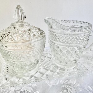 May include: A clear pressed glass sugar bowl with a lid, a matching creamer pitcher, and a rectangular tray. Each piece features a textured diamond pattern and scalloped edges. The sugar bowl has a pointed finial on the lid, while the creamer has a curved handle and a pouring spout.