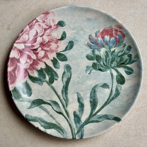 May include: A round ceramic plate featuring a light blue background with a large, detailed pink flower on the left and a smaller, multi-colored flower with blue, red, and green petals on the right, both connected by long, dark green stems and leaves.