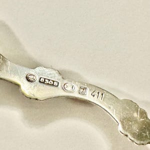 K&ouml;nnte beinhalten: Nahaufnahme eines silberfarbenen Metallobjekts mit verzierten Kanten. Das Objekt hat mehrere Markierungen, darunter "8365" und "411". Das Design ist detailliert und scheint Vintage oder antik zu sein.