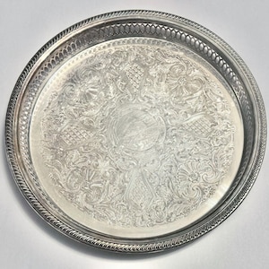 May include: A round silver-toned metal serving tray featuring an intricate embossed floral and scrollwork pattern across the center. The tray has a raised, openwork lattice border and a decorative twisted rope edge detail.