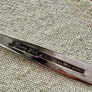 Może przedstawiać: Srebrzysty uchwyt sztućca ze stali nierdzewnej z wytłoczonym prostokątnym znakiem z napisem Towne Hall, STAINLESS, INOX oraz JAPAN. Metalowa powierzchnia ma odblaskowe wykończenie z wyraźnie widocznymi grawerowanymi literami.