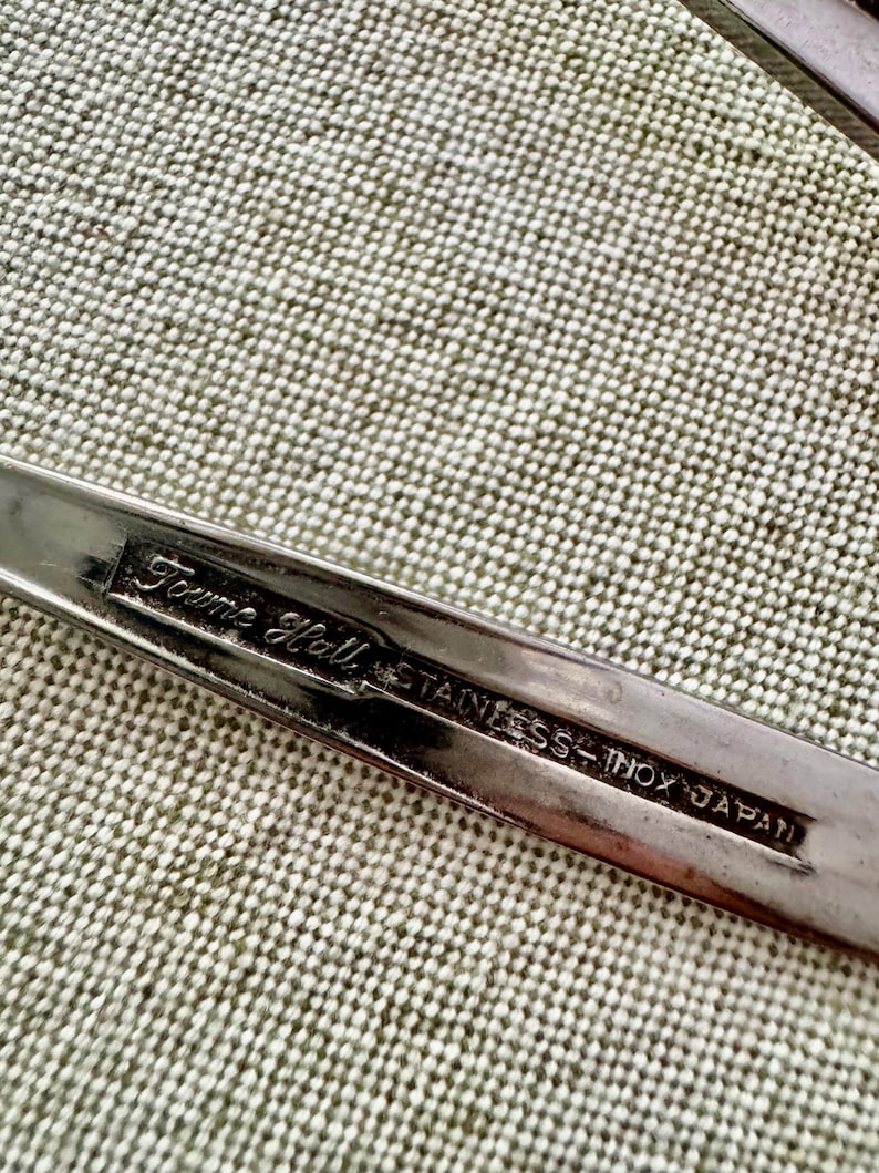 Może przedstawiać: Srebrzysty uchwyt sztućca ze stali nierdzewnej z wytłoczonym prostokątnym znakiem z napisem Towne Hall, STAINLESS, INOX oraz JAPAN. Metalowa powierzchnia ma odblaskowe wykończenie z wyraźnie widocznymi grawerowanymi literami.