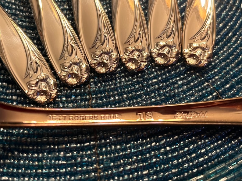 May include: Close-up of silver-colored flatware with floral designs. The spoons and forks have a detailed flower pattern at the base of the handle. The text "1847 Rogers Bros" is visible on the handle of one piece.