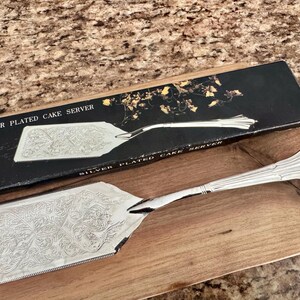 K&ouml;nnte beinhalten: Ein versilberter Tortenheber mit einer verzierten Blumen-Gravur auf der flachen Klinge und einem kannelierten Griff. Der Tortenheber wird zusammen mit seiner schwarzen Originalverpackung pr&auml;sentiert, die goldene Blumenakzente und den Text SILVER PLATED CAKE SERVER zeigt.