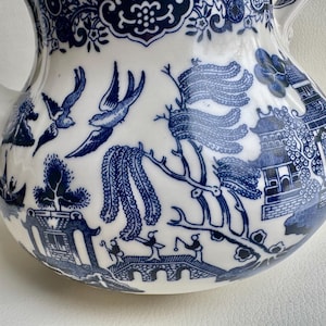 K&ouml;nnte beinhalten: Eine wei&szlig;e Teekanne mit einem blauen und wei&szlig;en Weidenmuster. Die Teekanne hat einen runden K&ouml;rper und einen Ausguss auf der linken Seite. Das Muster enth&auml;lt V&ouml;gel, B&auml;ume und eine Br&uuml;cke.