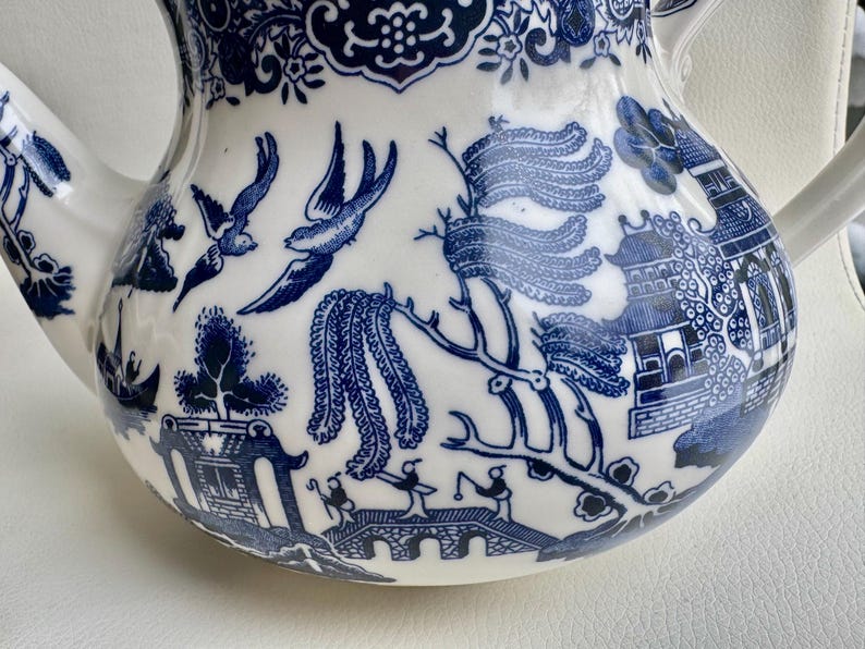 K&ouml;nnte beinhalten: Eine wei&szlig;e Teekanne mit einem blauen und wei&szlig;en Weidenmuster. Die Teekanne hat einen runden K&ouml;rper und einen Ausguss auf der linken Seite. Das Muster enth&auml;lt V&ouml;gel, B&auml;ume und eine Br&uuml;cke.