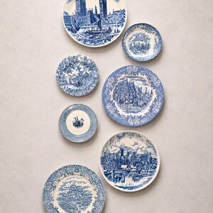May include: A collection of seven round ceramic plates featuring blue and white transferware patterns. The designs include detailed illustrations of historical buildings, landscapes, and architectural scenes, with varied floral and geometric borders.