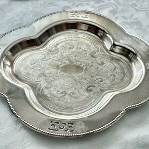 May include: A silver-colored, ornate serving tray with a quatrefoil shape. The tray features an embossed floral design in the center and decorative edging. The tray is likely made of metal and is suitable for serving food or displaying items.