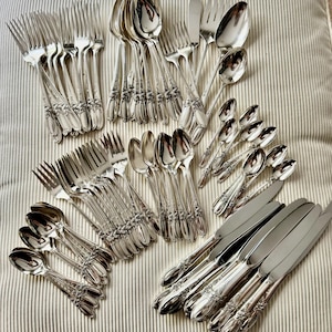 Op de afbeelding: Een assortiment vintage verzilverd bestek, waaronder vorken, lepels, messen en botermessen. Het zilverwerk heeft sierlijke bloemendetails op de handvatten. De opstelling staat op een gestreept, gebroken witte ondergrond.