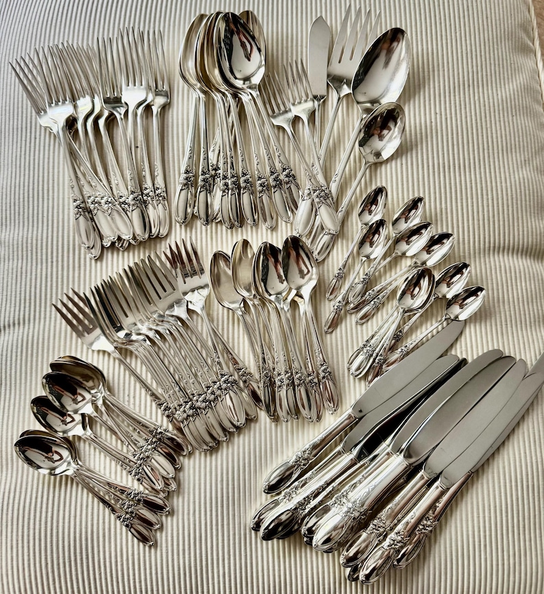 Op de afbeelding: Een assortiment vintage verzilverd bestek, waaronder vorken, lepels, messen en botermessen. Het zilverwerk heeft sierlijke bloemendetails op de handvatten. De opstelling staat op een gestreept, gebroken witte ondergrond.