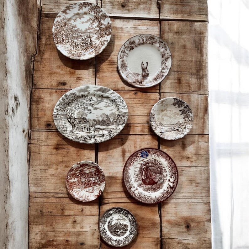 May include: A collection of seven ceramic plates and platters featuring brown and white transferware patterns. The designs include a rabbit, a turkey, and various landscape scenes with buildings and trees, arranged in a display on a wooden surface.