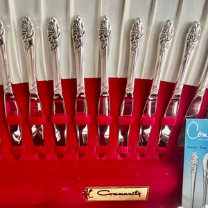 May include: A set of Community silverware knives arranged in a red velvet-lined case. The knives have a decorative floral design on the handles. The case also contains a brochure with the Community logo and information about the silverware patterns.