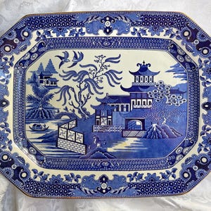 May include: An octagonal ceramic platter featuring a blue and white willow pattern. The design depicts a traditional landscape with pagodas, a bridge, a boat, and two birds in flight. The border is decorated with intricate geometric and floral motifs.