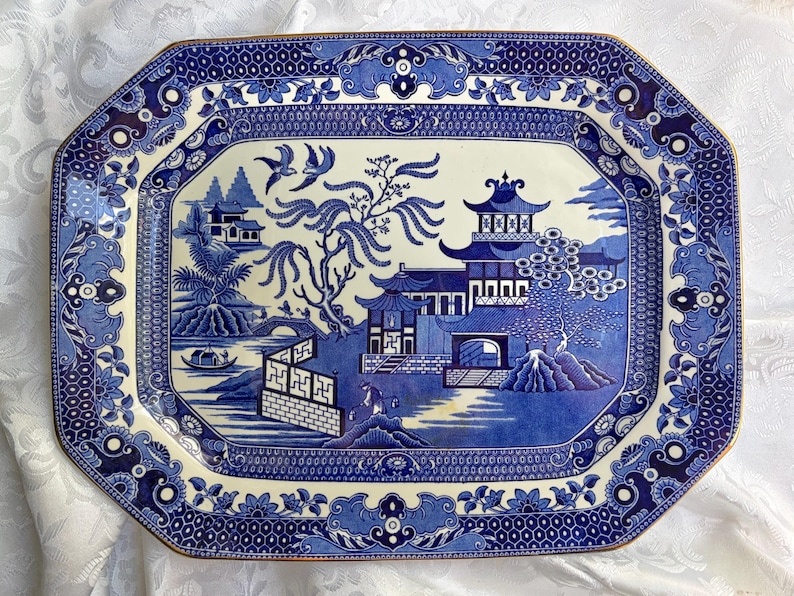 May include: An octagonal ceramic platter featuring a blue and white willow pattern. The design depicts a traditional landscape with pagodas, a bridge, a boat, and two birds in flight. The border is decorated with intricate geometric and floral motifs.