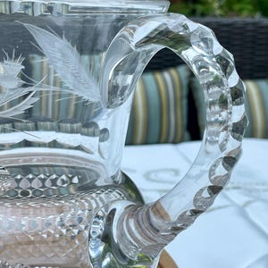 May include: A clear glass pitcher with a decorative handle and etched thistle design. The pitcher has a diamond-cut pattern on the base and a smooth rim. The handle is faceted, adding to the elegant aesthetic. Ideal for serving beverages.