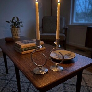 Peut inclure: Deux bougies allum&eacute;es, de couleur cr&egrave;me, dans des bougeoirs argent&eacute;s au design en spirale. Les bougeoirs sont pos&eacute;s sur une table basse en bois fonc&eacute;, &agrave; c&ocirc;t&eacute; de livres et d'un bol d&eacute;coratif. La sc&egrave;ne se d&eacute;roule dans une pi&egrave;ce faiblement &eacute;clair&eacute;e.