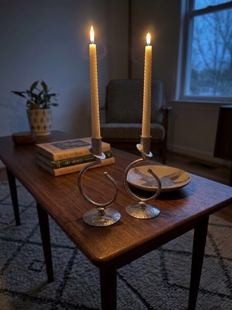 Peut inclure: Deux bougies allum&eacute;es, de couleur cr&egrave;me, dans des bougeoirs argent&eacute;s au design en spirale. Les bougeoirs sont pos&eacute;s sur une table basse en bois fonc&eacute;, &agrave; c&ocirc;t&eacute; de livres et d'un bol d&eacute;coratif. La sc&egrave;ne se d&eacute;roule dans une pi&egrave;ce faiblement &eacute;clair&eacute;e.