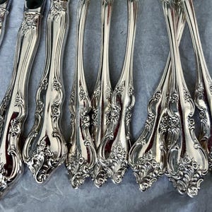 May include: Close-up of a collection of ornate silver forks and knives. The silverware features a detailed floral design on the handles. The polished metal reflects light, creating a bright and elegant appearance. The arrangement suggests a formal dining setting.