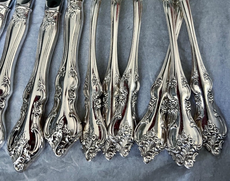 May include: Close-up of a collection of ornate silver forks and knives. The silverware features a detailed floral design on the handles. The polished metal reflects light, creating a bright and elegant appearance. The arrangement suggests a formal dining setting.
