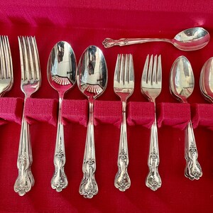 May include: A set of silver-toned flatware including forks, spoons, and a single horizontal spoon, all featuring ornate floral detailing on the handles. The utensils are arranged in a row within a red velvet-lined storage case.