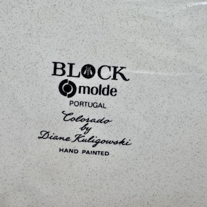 May include: A white ceramic surface with a speckled texture featuring black printed text that reads BLOCK, molde, PORTUGAL, Colorado by Diane Kuligowski, and HAND PAINTED. The text is arranged in a vertical stack with a bee icon in the word BLOCK.