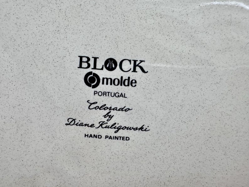 May include: A white ceramic surface with a speckled texture featuring black printed text that reads BLOCK, molde, PORTUGAL, Colorado by Diane Kuligowski, and HAND PAINTED. The text is arranged in a vertical stack with a bee icon in the word BLOCK.