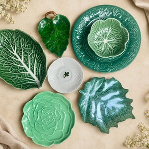 Puede incluir: Un conjunto de vajilla de cer&aacute;mica que incluye una fuente grande con forma de hoja de col verde, un plato peque&ntilde;o de hoja verde oscuro, un plato redondo verde con cuenco, un platillo blanco con motivo de hoja, un plato verde claro y un plato de hoja.