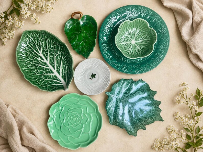 Puede incluir: Un conjunto de vajilla de cer&aacute;mica que incluye una fuente grande con forma de hoja de col verde, un plato peque&ntilde;o de hoja verde oscuro, un plato redondo verde con cuenco, un platillo blanco con motivo de hoja, un plato verde claro y un plato de hoja.
