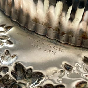 K&ouml;nnte beinhalten: Ein versilbertes Metallobjekt mit reflektierender Oberfl&auml;che, gepr&auml;gten Blumenmustern und vertikalen Rillen. Der Text "SILVER PLATED HONG KONG" ist deutlich in den glatten Mittelteil des metallischen Gegenstands eingraviert.