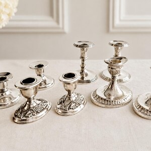 Pu&ograve; includere: Una collezione di sette portacandele in metallo color argento di varie altezze e design. Alcuni presentano basi lisce e lucide, mentre altri mostrano intricati motivi in rilievo di uva e foglie attorno alle basi circolari a piedistallo.