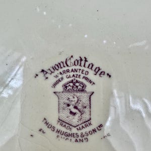 May include: A cream-coloured ceramic surface featuring a purple maker's mark. The text reads Avon Cottage, Warranted Under Glaze Print, Trade Mark, and Thos Hughes & Son Ltd England, accompanied by a shield emblem with a crown and a lion figure.