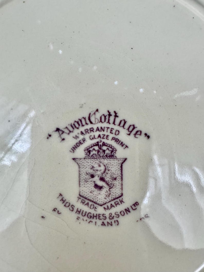 May include: A cream-coloured ceramic surface featuring a purple maker's mark. The text reads Avon Cottage, Warranted Under Glaze Print, Trade Mark, and Thos Hughes & Son Ltd England, accompanied by a shield emblem with a crown and a lion figure.