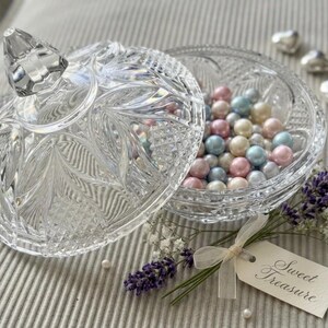 May include: A clear, pressed glass sweet dish with a faceted lid, filled with multicoloured pastel beads. A small tag tied with a cream ribbon reads "Sweet Treasure", accompanied by sprigs of purple lavender and white baby's breath flowers.