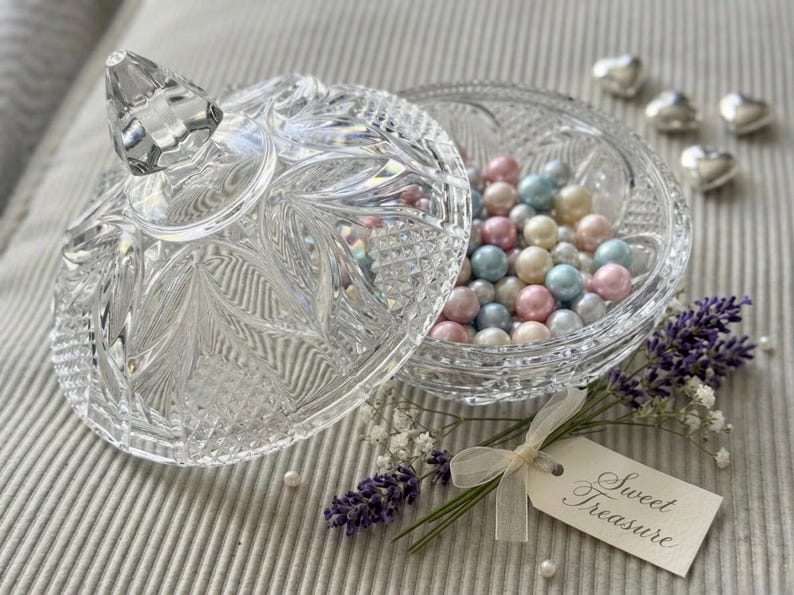 May include: A clear, pressed glass sweet dish with a faceted lid, filled with multicoloured pastel beads. A small tag tied with a cream ribbon reads "Sweet Treasure", accompanied by sprigs of purple lavender and white baby's breath flowers.